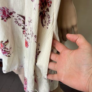Op de afbeelding: Cr&egrave;mekleurige top met bloemenprint en een split aan de zijkant. De stof heeft een getextureerd oppervlak en is versierd met bordeauxrode en roze bloempatronen. De top is mouwloos en lijkt van licht materiaal te zijn.
