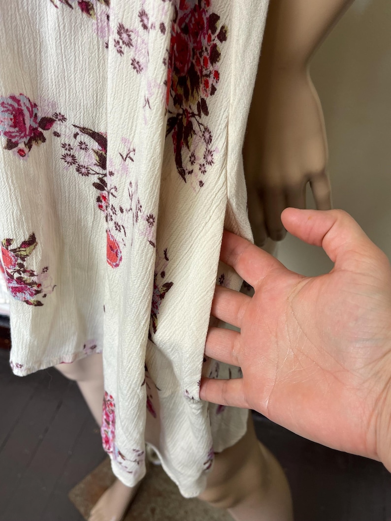 Op de afbeelding: Cr&egrave;mekleurige top met bloemenprint en een split aan de zijkant. De stof heeft een getextureerd oppervlak en is versierd met bordeauxrode en roze bloempatronen. De top is mouwloos en lijkt van licht materiaal te zijn.