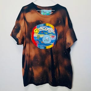 Op de afbeelding: Zwart en bruin tie-dye t-shirt met een cirkelvormige patch met een lachend gezicht, zeedieren en een stripfiguur. De patch is omrand met een regenboogkleurige rand. Het shirt hangt aan een witte hanger.
