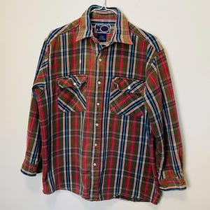 May include: A long-sleeved plaid shirt in shades of olive green, red, blue, and white. The shirt has two chest pockets, a button-down front, and a collared neckline. The shirt is hanging on a white hanger.