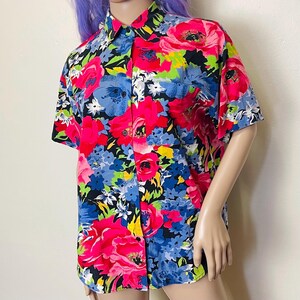 May include: Short-sleeved button-down shirt with a vibrant floral pattern. The shirt features a colorful design with large pink, blue, and yellow flowers against a black background. The shirt has a classic collar and two chest pockets.