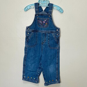 May include: Blue denim overalls with a butterfly design embroidered on the chest pocket. The overalls have adjustable straps and decorative stitching. Colorful heart-shaped embroidery accents the bottom hem. The overalls are hanging on a light blue hanger.