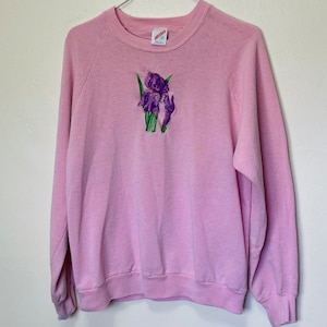 Puede incluir: Una sudadera rosa con un diseño de flor bordada en morado y verde.