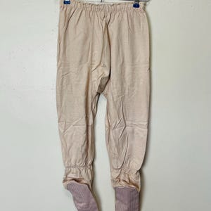 May include: Tan footed pajamas hanging on a metal hanger. The pajamas are a solid tan color with ribbed cuffs at the ankles and feet. The feet are a darker shade of tan with vertical stripes.