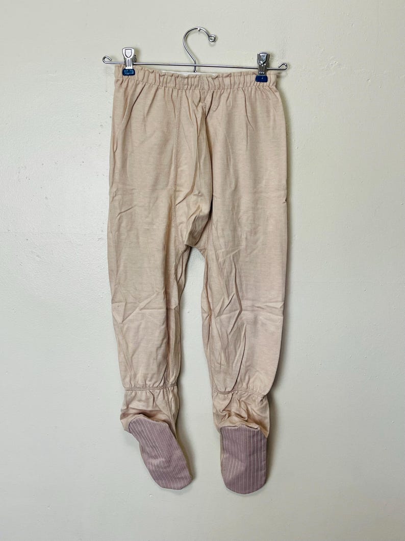 May include: Tan footed pajamas hanging on a metal hanger. The pajamas are a solid tan color with ribbed cuffs at the ankles and feet. The feet are a darker shade of tan with vertical stripes.