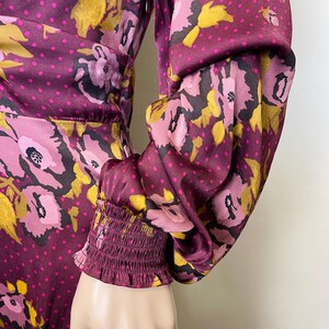May include: A long-sleeved dress with a floral print in shades of pink, purple, and gold. The dress has a fitted bodice and a flowing skirt. The sleeves are gathered at the wrist with an elastic band.