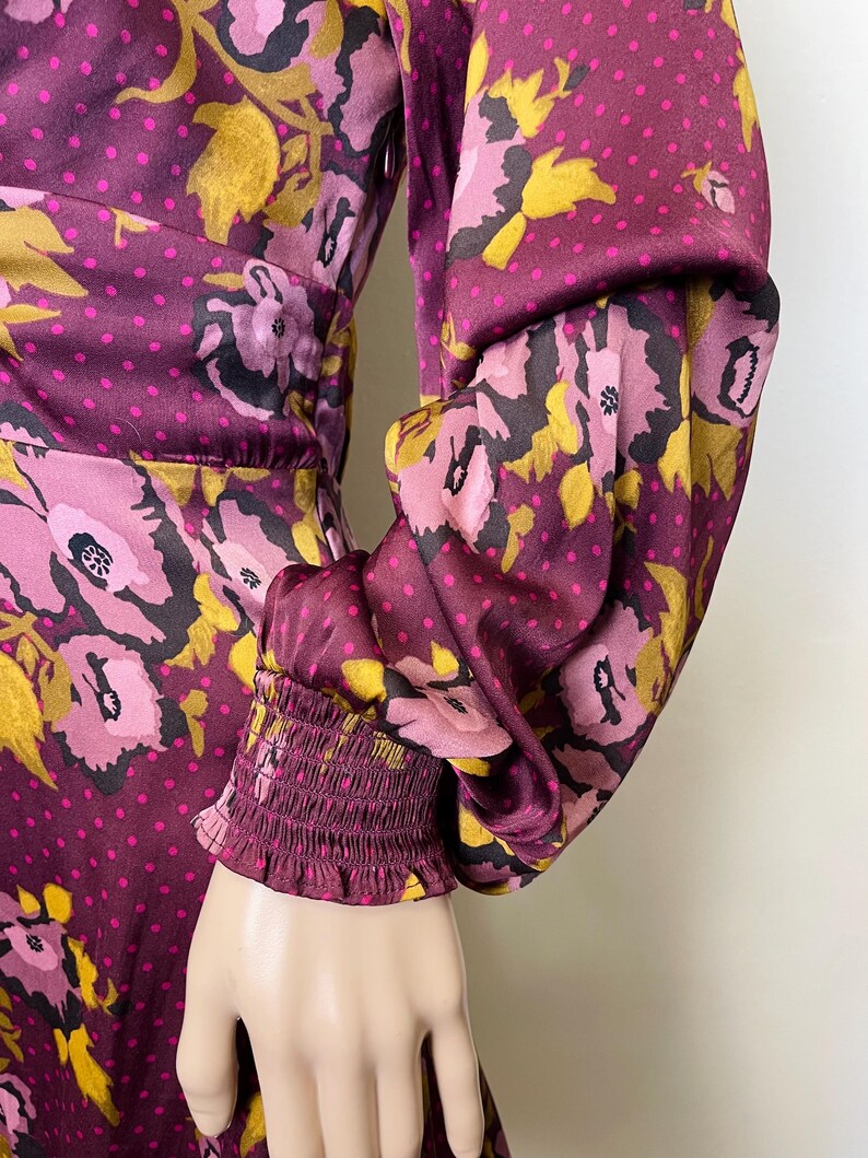 May include: A long-sleeved dress with a floral print in shades of pink, purple, and gold. The dress has a fitted bodice and a flowing skirt. The sleeves are gathered at the wrist with an elastic band.