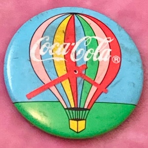 Puede incluir: Un pin vintage de Coca-Cola con un globo aerostático con un diseño de arcoíris. El globo está volando sobre un campo verde y el texto "Coca-Cola" está escrito en blanco en el globo.