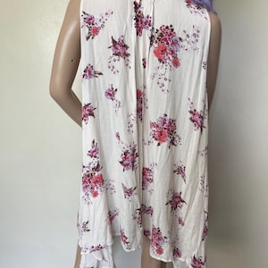 Op de afbeelding: Een mouwloze cr&egrave;mekleurige jurk met een bloemenpatroon in roze en rode tinten. De jurk heeft een asymmetrische zoom en een V-hals. De bloemenprint is verspreid over de stof.