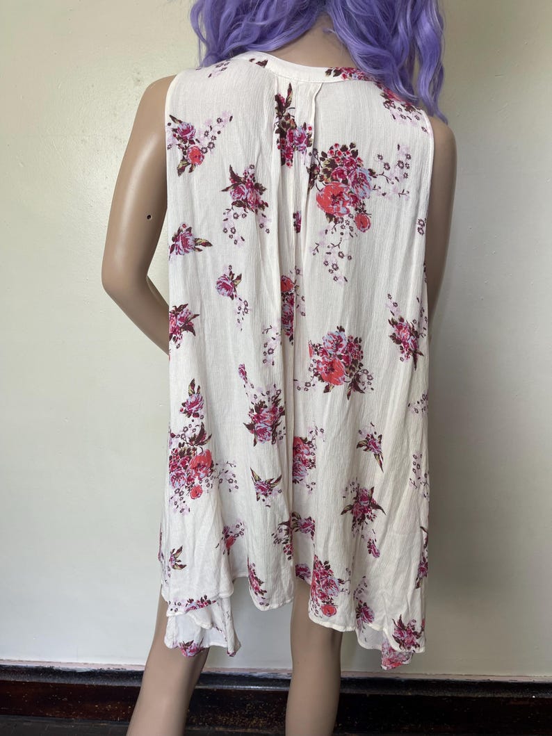 Op de afbeelding: Een mouwloze cr&egrave;mekleurige jurk met een bloemenpatroon in roze en rode tinten. De jurk heeft een asymmetrische zoom en een V-hals. De bloemenprint is verspreid over de stof.