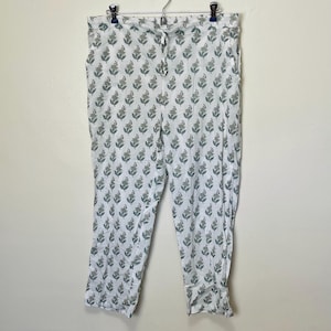 May include: White pajama pants with a repeating pattern of green floral sprigs. The pants have a drawstring waist and pockets. The fabric appears to be lightweight and suitable for sleepwear or lounging. The pants are hanging on a metal hanger.