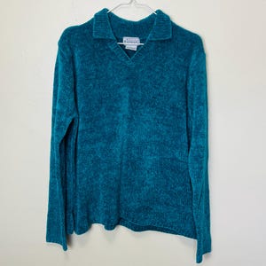 May include: A teal blue fuzzy sweater with a v-neck and a collared neckline. The sweater has a small white tag with the text "Betty & Frank" on it.