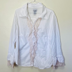 Puede incluir: Camisa blanca abotonada con mangas largas y un acento de encaje rosa claro con volantes en la parte delantera. Los puños también presentan un ribete de encaje a juego. La camisa tiene cuello y la marca "unique" es visible en la etiqueta.