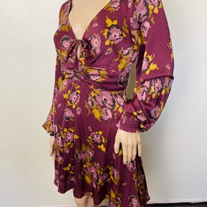 May include: A burgundy floral print dress with a V-neck, long sleeves, and a cinched waist. The dress has a tie detail at the neckline and a ruffled hem.