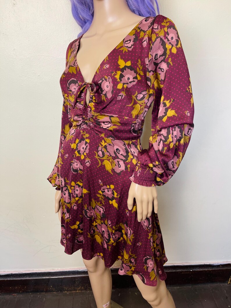 May include: A burgundy floral print dress with a V-neck, long sleeves, and a cinched waist. The dress has a tie detail at the neckline and a ruffled hem.