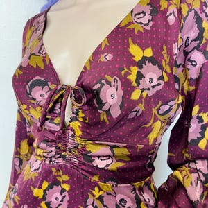 May include: A long-sleeved, purple floral print dress with a gathered neckline and a tie detail. The dress has a polka dot pattern on a dark purple background.