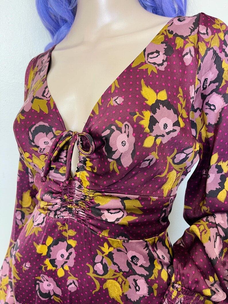 May include: A long-sleeved, purple floral print dress with a gathered neckline and a tie detail. The dress has a polka dot pattern on a dark purple background.