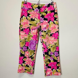 May include: Cropped trousers with a vibrant floral pattern. The trousers feature a black background with large pink, purple, and peach-coloured flowers and green leaves. The trousers are hanging on a white background.
