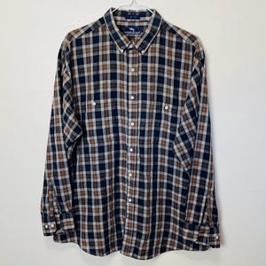 Puede incluir: Una camisa de manga larga a cuadros en marrón, negro y blanco. La camisa tiene un cuello estándar y un cierre de botones en la parte delantera. La etiqueta de la marca dice "Steeplechase".