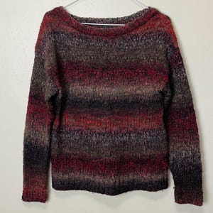 May include: A multi-colored sweater with a boat neck. The sweater features horizontal stripes in shades of red, brown, black, and gray. The long-sleeved sweater is hanging on a white hanger.
