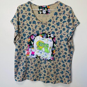 Könnte beinhalten: Ein beiges T-Shirt mit kurzen Ärmeln und einem Paisley-Muster. Ein farbenfroher Patch mit zwei Comic-Löwen und dem Text "I love you something fierce" ist auf der Vorderseite des Shirts aufgenäht.