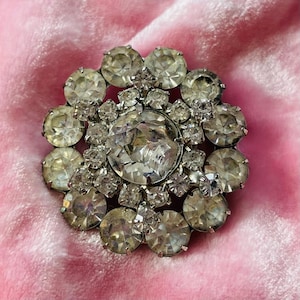 Puede incluir: Un broche de plata con un gran strass transparente rodeado de strass más pequeños.