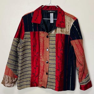 May include: A patchwork jacket with a collared neckline and button-up closure. The jacket features a mix of red, black, gold, and tan fabrics with floral embroidery and paisley patterns. The sleeves are long and the jacket is hanging on a hanger.
