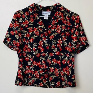 May include: A short-sleeved black button-down shirt with a floral pattern. The pattern features red flowers and beige leaves. The shirt has a collar and is hanging on a white hanger.