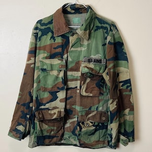 Könnte beinhalten: Eine langärmelige Camouflage-Jacke mit Waldmuster in Grün-, Braun- und Schwarztönen. Die Jacke verfügt über vier Vordertaschen mit geknöpften Klappen, einen Kragen und einen schwarzen, gestickten U.S. ARMY-Aufnäher über der Brusttasche.