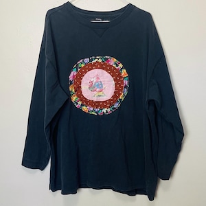 May include: A dark navy blue long-sleeve sweatshirt with a crew neck. The sweatshirt features a circular appliqué with a pink center, a brown border, and a floral pattern. The appliqué depicts a cartoon character holding flowers.