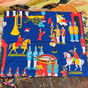 Könnte beinhalten: Ein blauer Stoff mit einem sich wiederholenden Muster aus Marschkapellenfiguren, Kanonen und Flaggen. Die Figuren tragen rote, gelbe und blaue Uniformen. Eine große gelbe Trompete ist ebenfalls im Design enthalten.