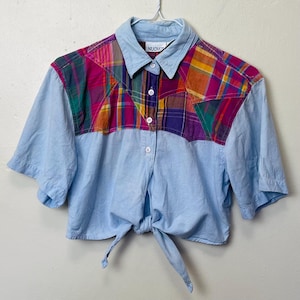 May include: A cropped button-down shirt with short sleeves. The shirt features a light blue body and a colorful plaid yoke in shades of pink, purple, green, and orange. The shirt has a tie closure at the waist and a collar.