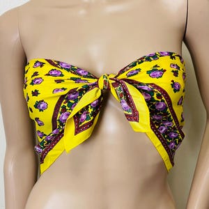 May include: A yellow scarf tied in a knot, featuring a floral pattern with purple flowers and green leaves. The scarf has a red and brown border. The scarf is wrapped around a mannequin.