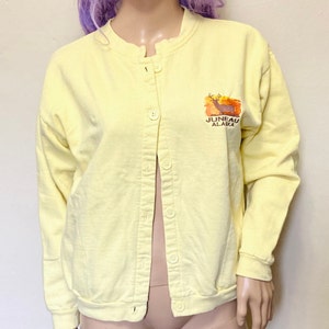 Peut inclure: Un sweat-shirt jaune à boutons avec un motif de cerf et le texte "Juneau Alaska".