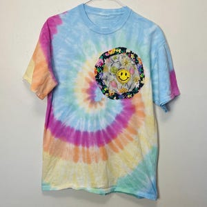 May include: A tie-dye t-shirt with a spiral pattern in shades of blue, pink, yellow, and orange. A circular patch featuring a yellow smiley face and floral design is on the chest. The shirt has short sleeves and a crew neck.