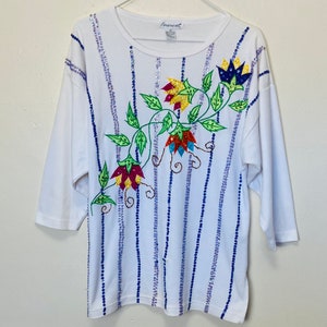 May include: White long-sleeved shirt with a floral design embroidered with sequins. The flowers are in shades of red, yellow, blue, and green. The shirt has a vertical striped pattern of blue and purple sequins.