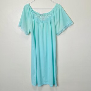 May include: A light turquoise nightgown featuring a wide scoop neckline with white floral embroidery and scalloped edges. The garment has short, loose sleeves finished with delicate white lace trim along the cuffs and a simple, straight hemline.