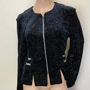 Puede incluir: Una chaqueta de terciopelo negro con purpurina plateada en todo. La chaqueta tiene una cremallera y dos hebillas plateadas decorativas a los lados.