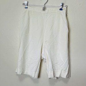 May include: Cream-colored, high-waisted shorts hanging on a metal hanger. The shorts have a tailored design with a button closure and a zipper. The fabric appears to be a lightweight material, suitable for warmer weather. The shorts are hanging against a neutral-colored wall.