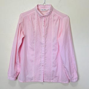 Puede incluir: Una camisa rosa de manga larga con botones, cuello Peter Pan y ribete de encaje. La camisa tiene un frente plisado y la etiqueta "Rowan & Buckley" es visible en el cuello.