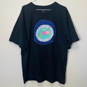 Puede incluir: Camiseta negra con una aplicación floral. La aplicación presenta un diseño circular con un borde azul punteado, un centro azul claro y flores rosas, blancas y amarillas. La camiseta tiene mangas cortas y cuello redondo.