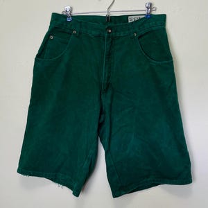 Peut inclure: Shorts en jean vert émeraude avec une conception classique à cinq poches. Les shorts ont une fermeture à bouton et fermeture éclair, des passants de ceinture et un aspect légèrement usé à l'ourlet. Les shorts sont suspendus à un cintre en métal.