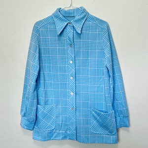 Puede incluir: Una camisa de color azul claro con cuello y un estampado de cuadros blanco y azul claro. La camisa tiene un cierre de botones en la parte delantera y dos bolsillos.