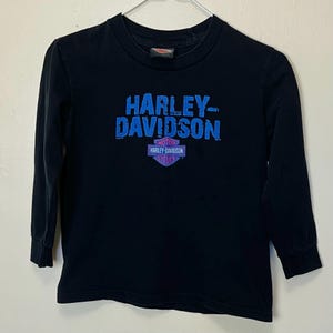 May include: Black long-sleeved shirt with the Harley-Davidson logo in blue and purple. The logo includes the text "HARLEY-DAVIDSON" and "MOTOR HARLEY-DAVIDSON CYCLES". The shirt is hanging on a white hanger.