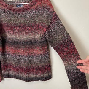 May include: A multi-colored sweater with a crew neck. The sweater features a blend of red, brown, and gray hues. The brand label "CHAPS" is visible on the upper back. The sweater is made of a knitted material.