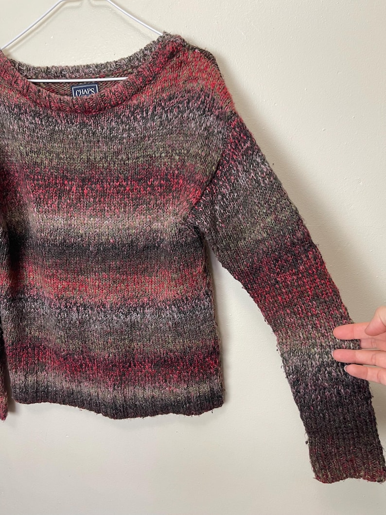 May include: A multi-colored sweater with a crew neck. The sweater features a blend of red, brown, and gray hues. The brand label "CHAPS" is visible on the upper back. The sweater is made of a knitted material.