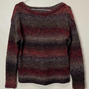 May include: A multi-colored sweater with a boat neck. The sweater features horizontal stripes in shades of red, brown, and gray. The long-sleeved sweater is hanging on a white hanger against a white background.