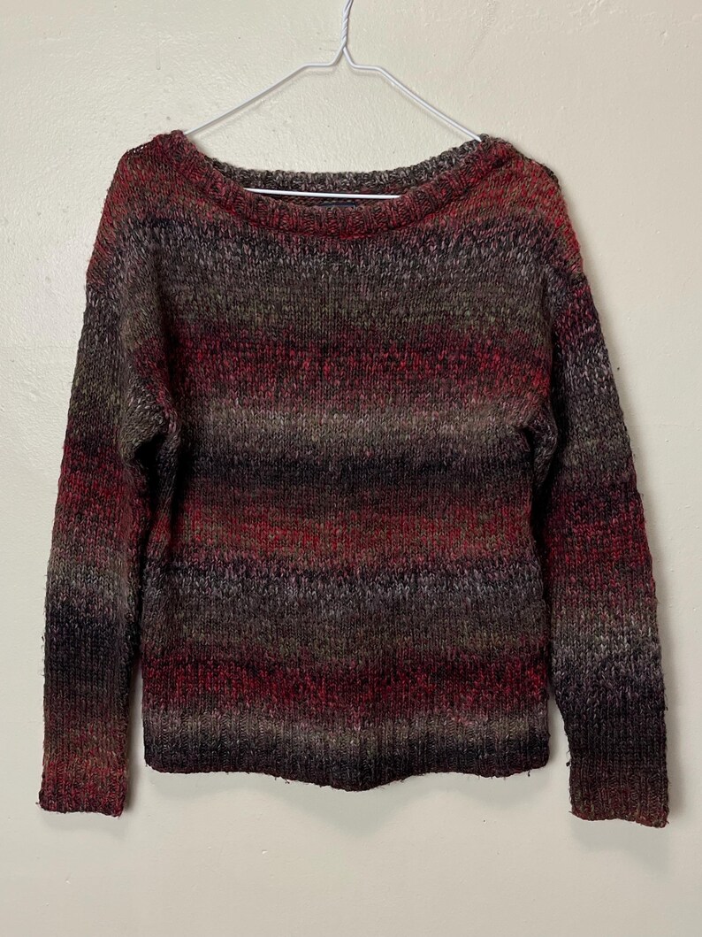 May include: A multi-colored sweater with a boat neck. The sweater features horizontal stripes in shades of red, brown, and gray. The long-sleeved sweater is hanging on a white hanger against a white background.