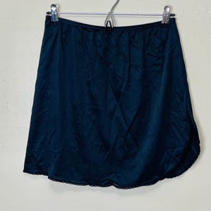 May include: A black slip skirt hanging on a metal hanger. The skirt is made of a smooth, shiny fabric and has a scalloped lace trim along the hemline. The skirt is a-line in shape.