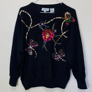 May include: Black sweater with colourful floral and butterfly sequins. The sweater has a crew neck and long sleeves. The brand label reads "Alfani".